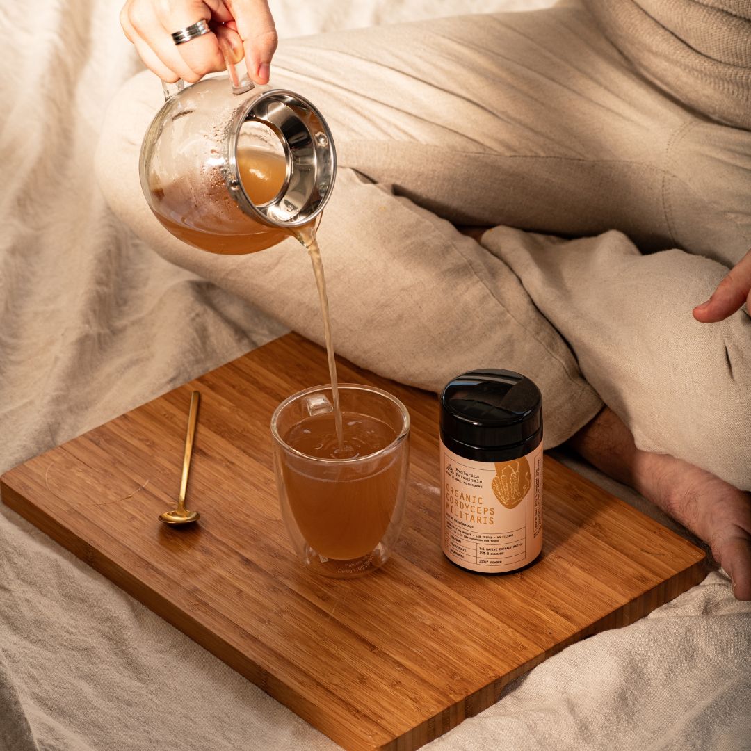 A 100g jar of Organic Cordyceps Militaris next to a glass cup with liquid in it being poured into from a glass tea pourer