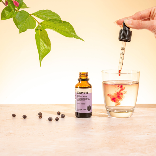 A 30ml dropper bottle of SoWell Elderberry liquid on a table next to a handle dropping its liquid into a glass of water