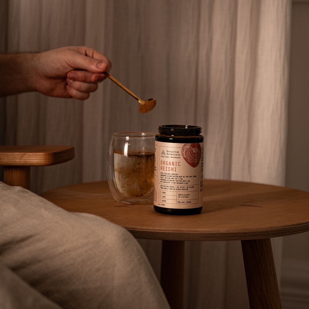 A closeup of a person spooning brown powder into a glass cup of water next to a 90g jar of Organic Reishi