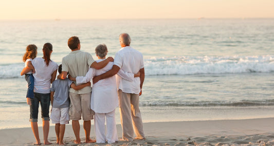 A group of 5 people of various ages standing inline on a beach staring off into the water watching a sunset. the