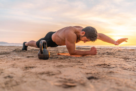 A man doing a one armed plank on a beach with a sunset behind him.  