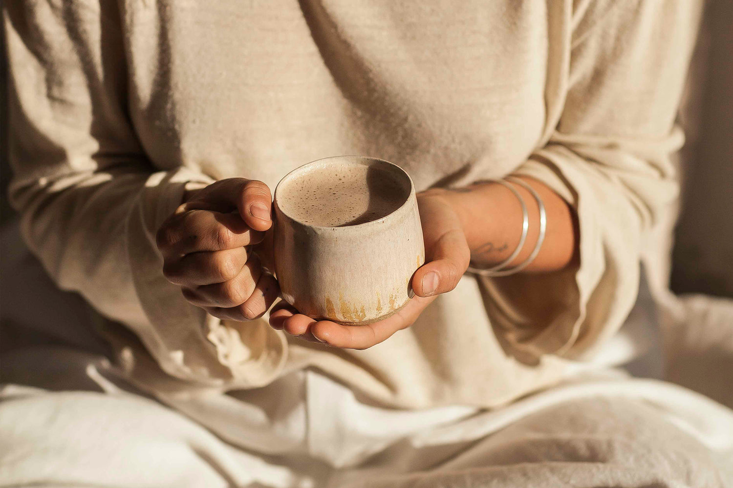 A close up shot of a woman from the torso down, wearing linen clothes and holding a cup of warm liquid in a coffee mug