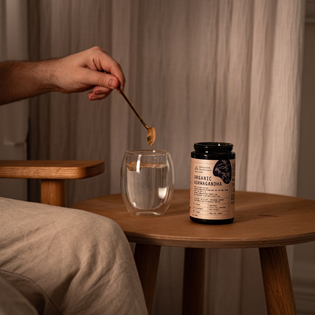 A jar of Organic Ashwagandha on a table next to a glass of water with a person's hand spooning Ashwagandha into the liquid