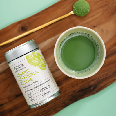Jar of Organic Ceremonial Matcha next to a mug of matcha drink and a spoon lying on a wooden board