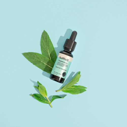 A wide shot of a 30ml dropper bottle of Sowell Peppermint tincture surrounded by peppermint leaves on a blue background